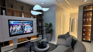 A modern living room with a large flat-screen TV, a curved grey sofa, a coffee table, and a bookshelf. - Phaluxe Shortlets (Abuja)
