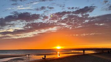 Escena de playa con el sol poniéndose en el horizonte, gente caminando por la orilla y un perro.