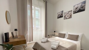 Quarto de hotel com uma cama, escrivaninha com laptop, cadeira, mesinha de cabeceira com luminária e planta em vaso.