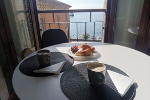 Een ronde tafel met een witte tafelkleed, twee zwarte placemats, een kop koffie, een croissant, en een bord met gebak en bessen.