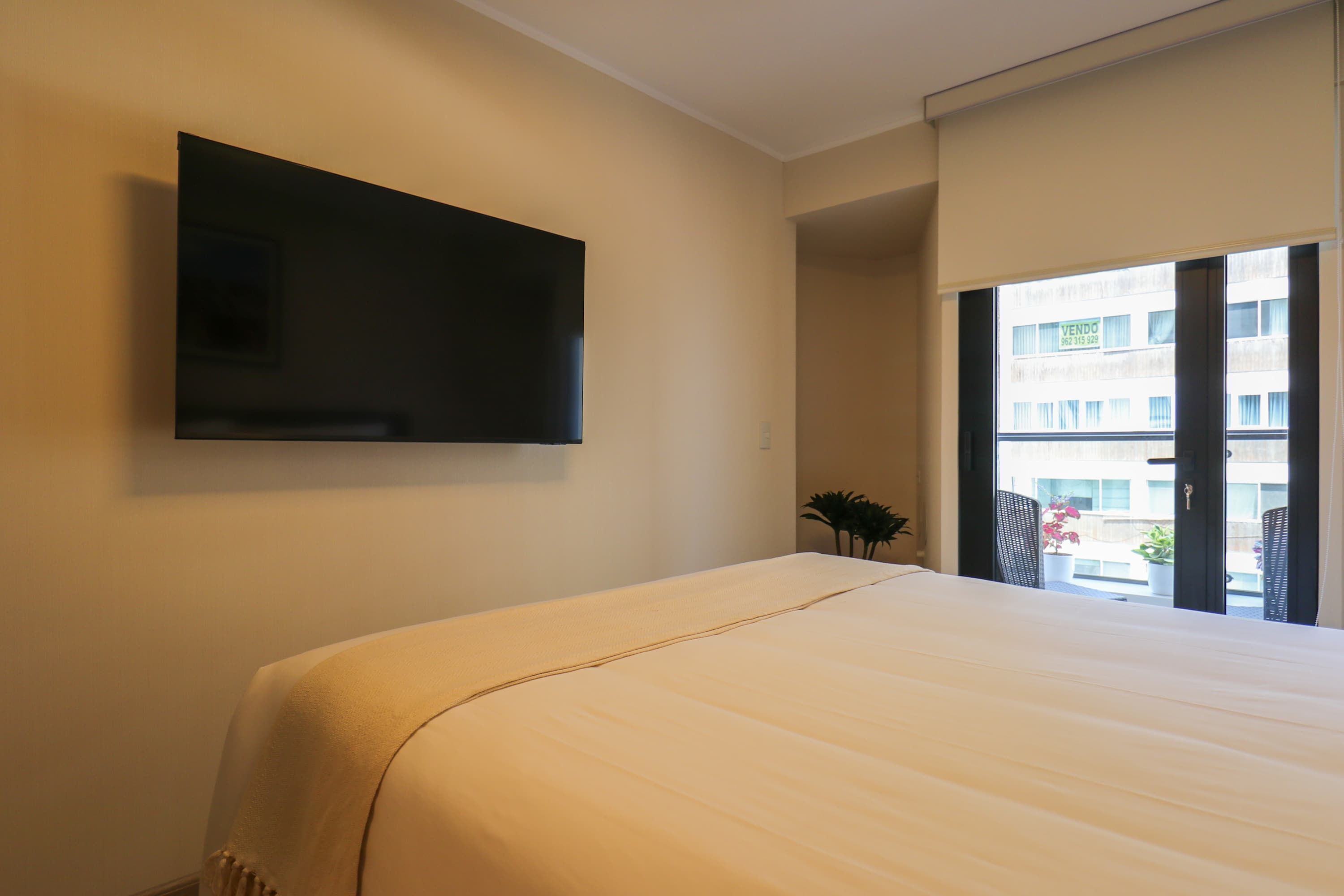 Hotellihuone, jossa on sänky, seinälle asennettu televisio ja parveke, jolta on näkymä rakennuksiin.