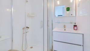 Un baño moderno con ducha envuelta en vidrio, tocador blanco con espejo y un letrero de emergencia verde.