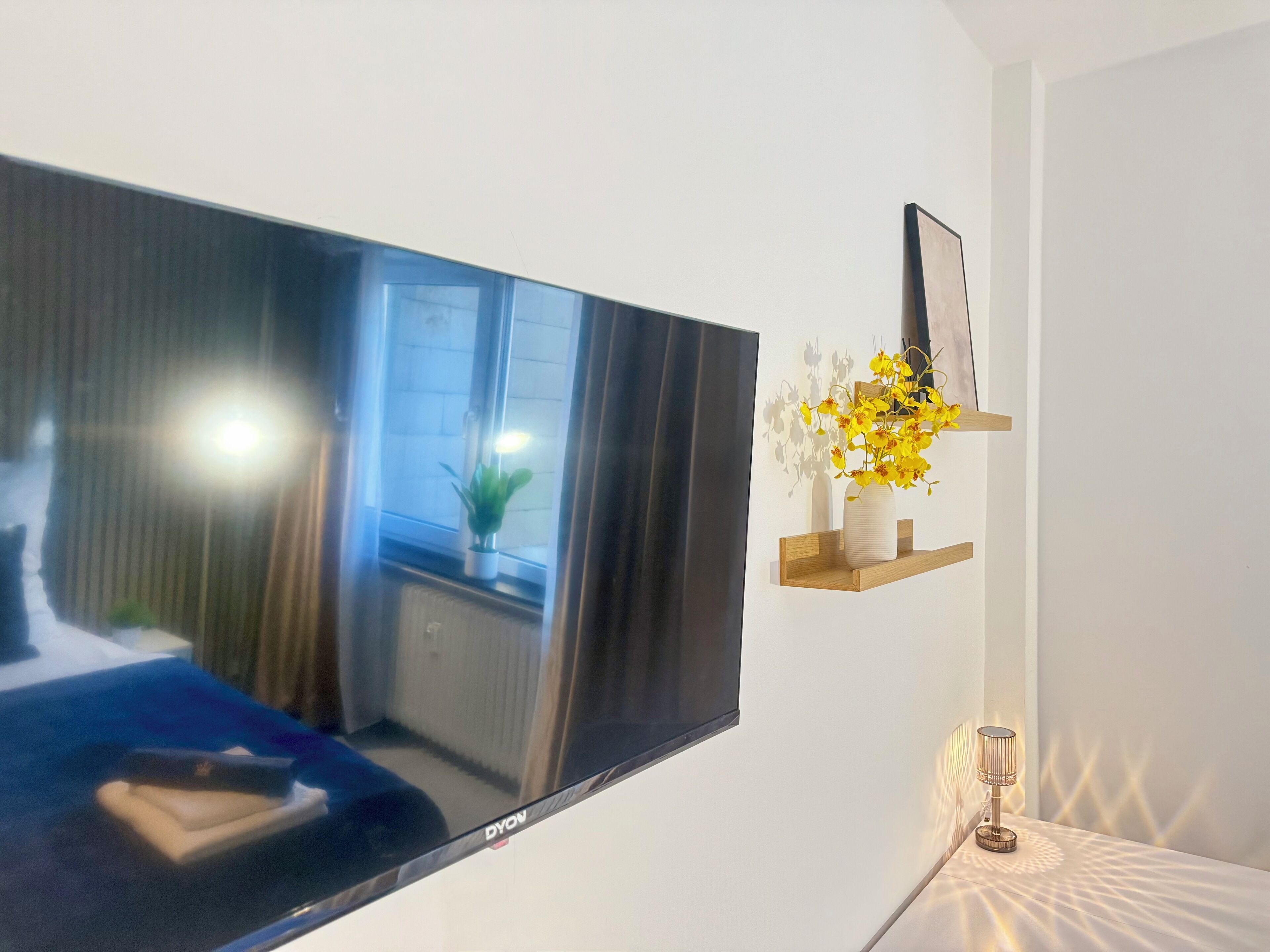 A modern living room with a flat-screen TV mounted on the wall, a wooden shelf with a vase of yellow flowers, and a lamp on a bedside table.
