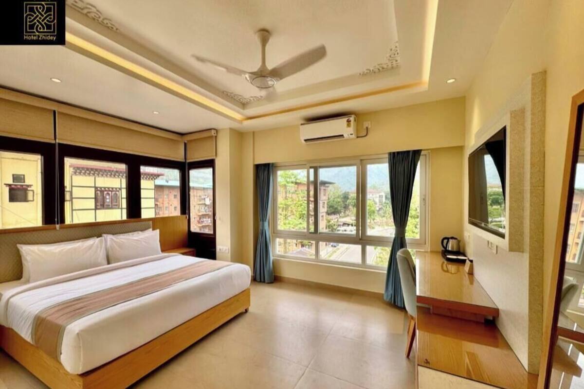 A hotel room with a bed, a TV, a ceiling fan, and a window with curtains.