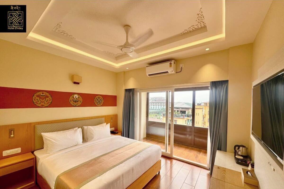 A hotel room with a large bed, a ceiling fan, an air conditioning unit, a desk with a chair, and a view of the city through the window.