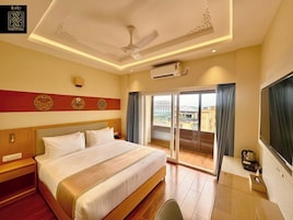 A hotel room with a large bed, a ceiling fan, an air conditioning unit, a desk with a chair, and a view of the city through the window.