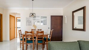 Un coin repas avec une table et des chaises en bois, un luminaire suspendu, des œuvres d’art encadrées au mur et un canapé vert au premier plan.