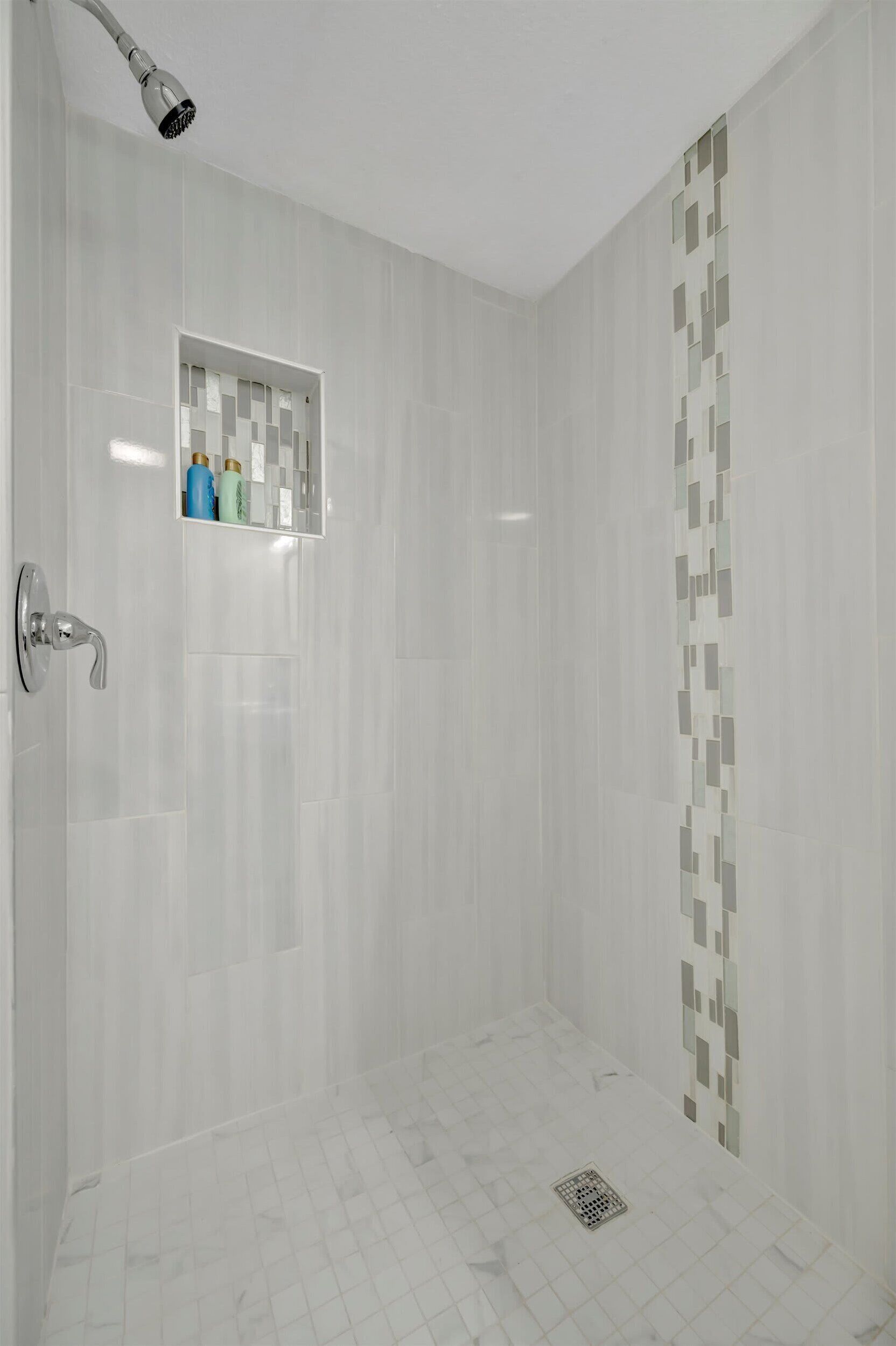 Een moderne douche met een handdouche, een klein plankje met toiletartikelen en een betegelde vloer met afvoer.