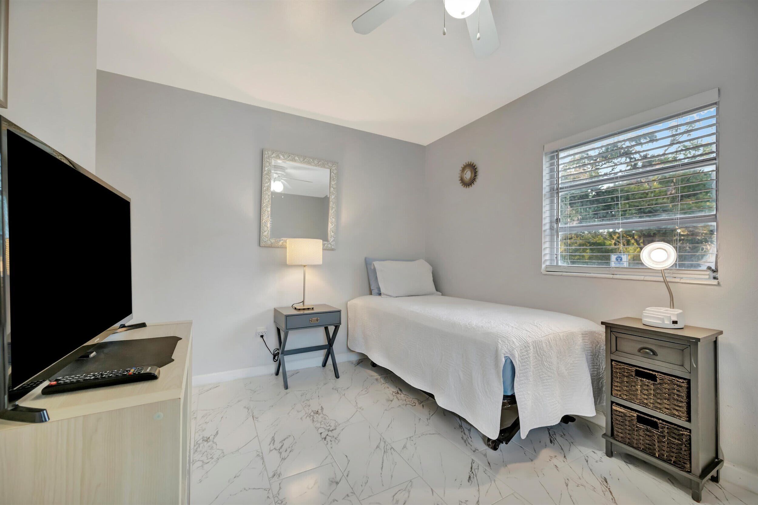 A bedroom with a bed, a TV, a lamp, a window with blinds, and a ceiling fan.