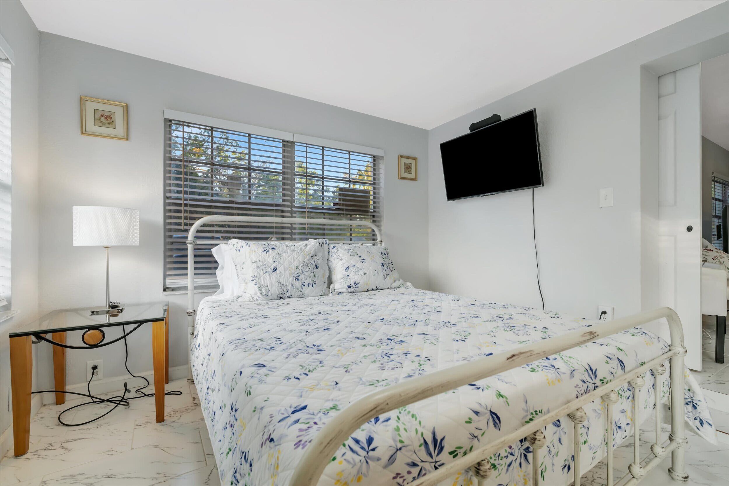 A bedroom with a bed, bedside table, lamp, TV, and window with blinds.