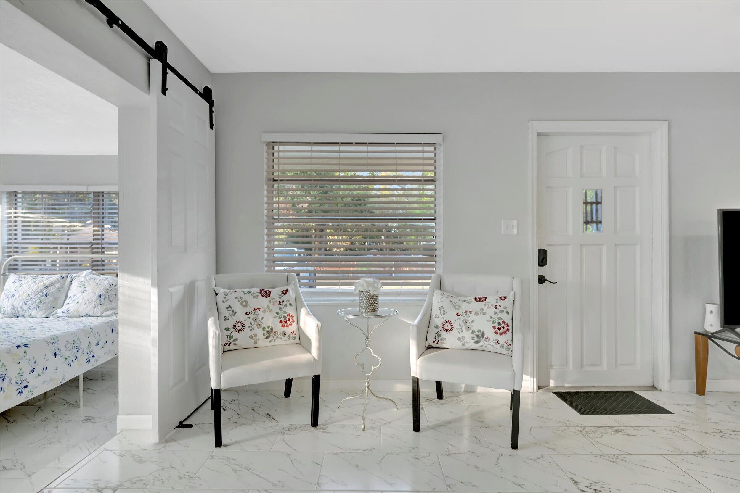 Una habitación moderna con una cama, dos sillas blancas, una mesita y una puerta.