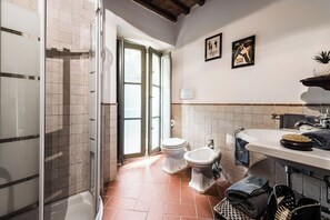 A bathroom with a shower, toilet, bidet, and sink. There are two doors leading to an outdoor area.