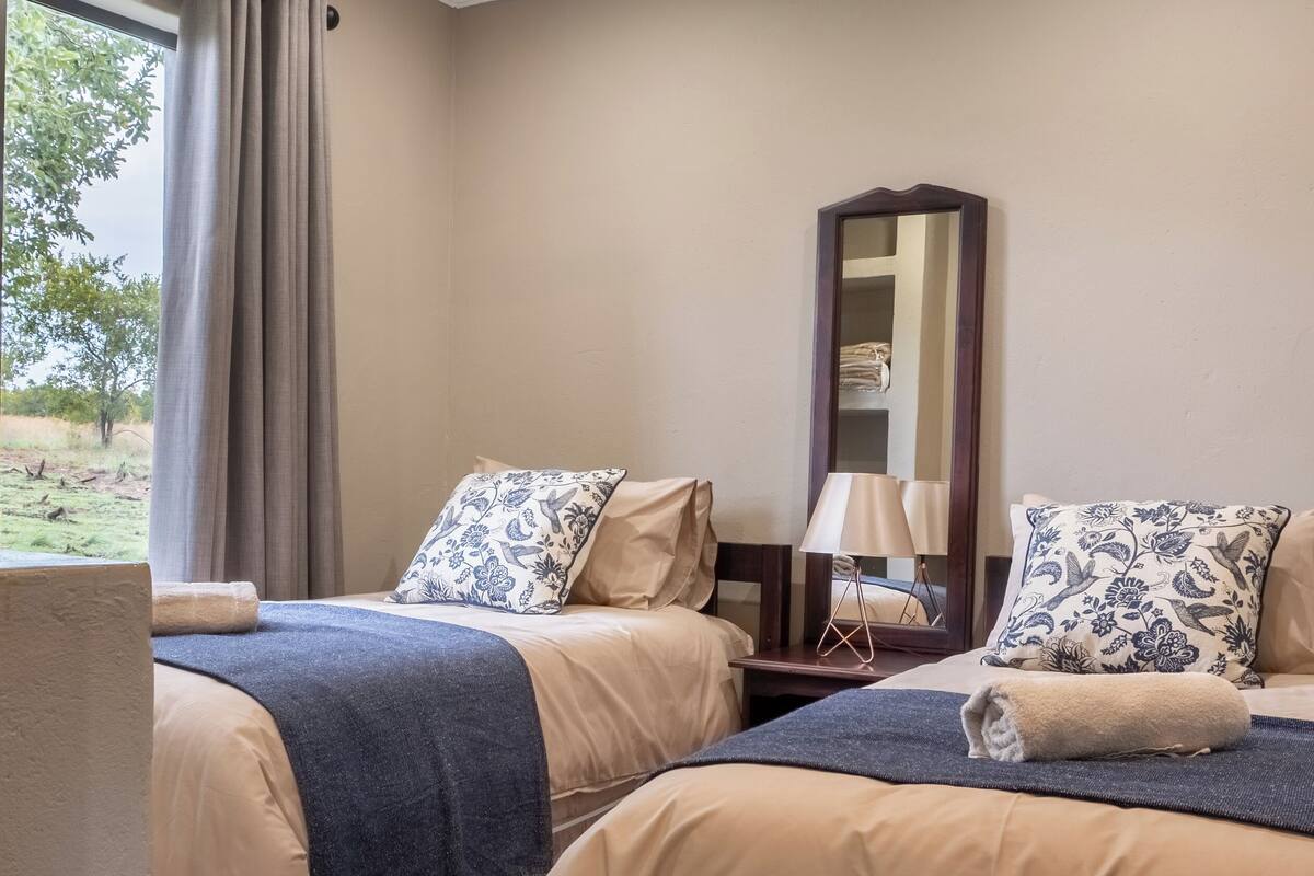 Ein Hotelzimmer mit zwei Betten, einem Deckenventilator, einem großen Fenster mit Vorhängen, einem Spiegel und einer Lampe.
