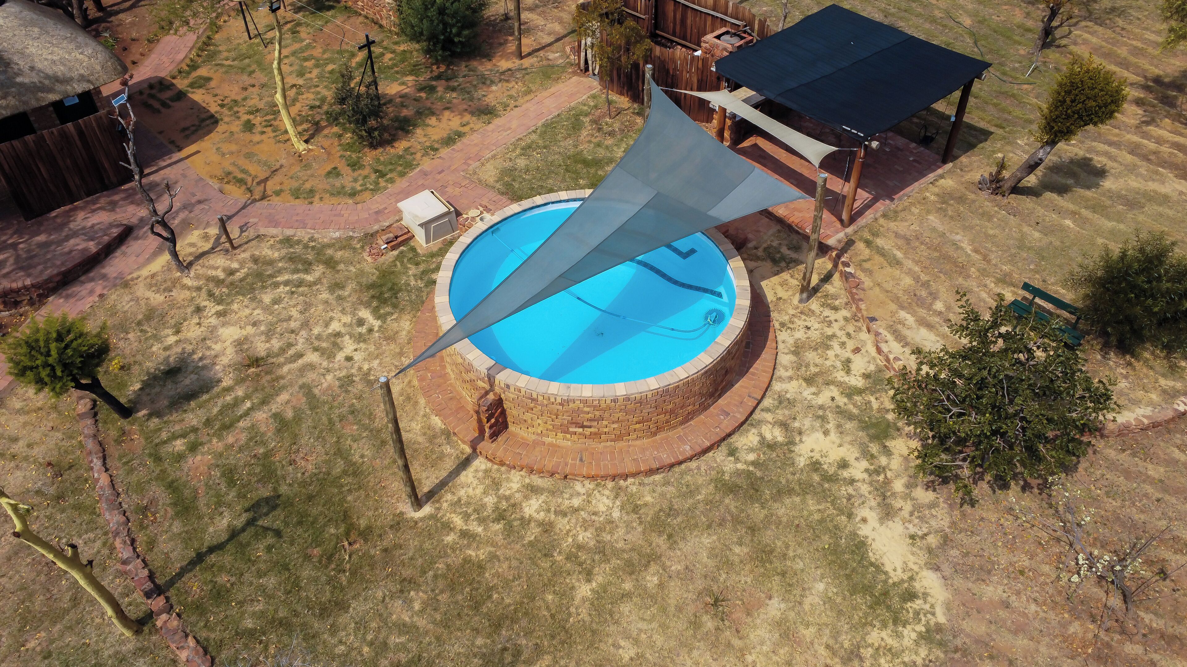 An aerial view of a backyard with a swimming pool, surrounded by a brick patio, a wooden deck, and a fenced area with a pergola.