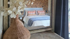 Un miroir reflétant une chambre à coucher avec un mur en pierre, des poutres en bois au plafond, un lit avec des coussins à motifs et un vase tressé contenant des fleurs blanches.