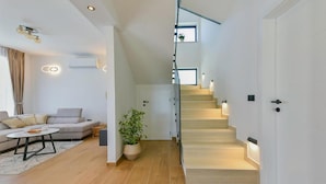 Een moderne woonkamer met een bank, salontafel en een potplant. Een trap leidt naar een tweede verdieping met een deur.