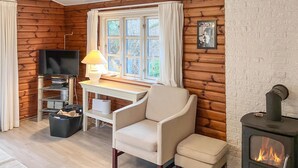 Een gezellig chaletinterieur met een houten wand, een televisie, een bureau met lamp, een stoel, een klein tafeltje, een prullenbak en een houtkachel.