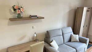 A modern living room with a grey sofa, a wooden desk with a chair, a small shelf with books and a vase of flowers, and a wardrobe. - Cozy Corner Ljubljana - Modern Urban Retreat (Ljubljana)