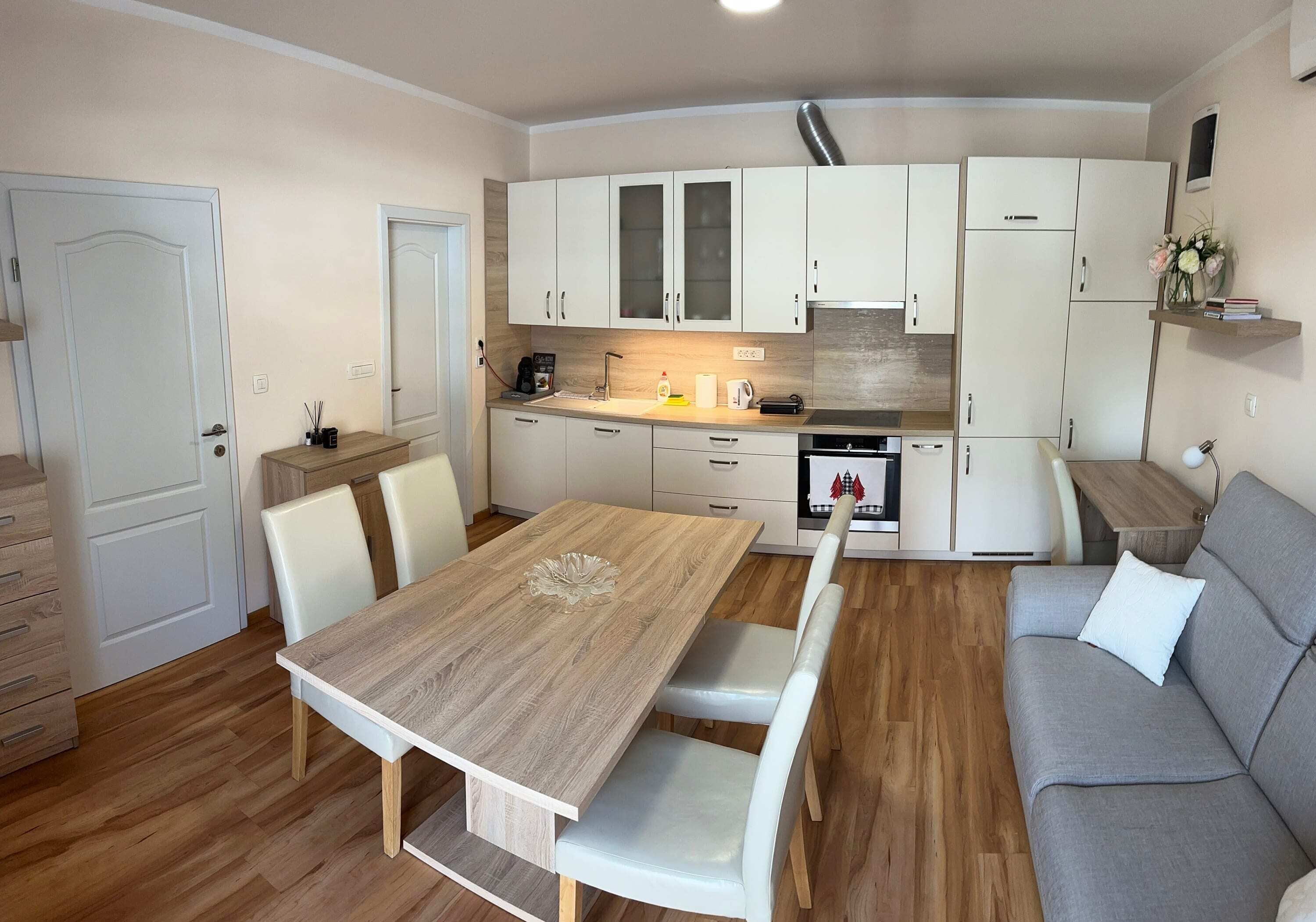 Una cocina funcional con armarios blancos, una mesa de madera con sillas y un sofá gris.