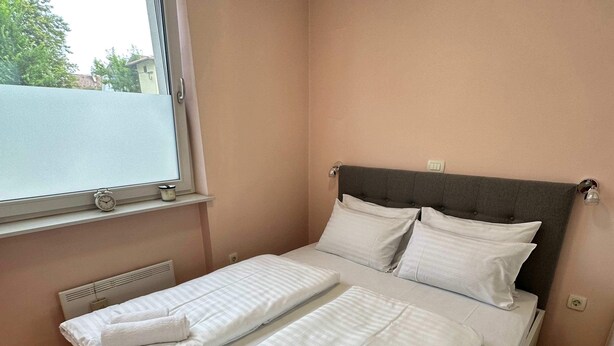 A neatly made bed with white linens and rolled towels, a window with blinds, and a radiator under the headboard. - Cozy Corner Ljubljana - Modern Urban Retreat (Ljubljana)