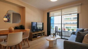 Una sala de estar moderna con un televisor de pantalla plana, una mesa de comedor con sillas, un sofá y una mesa de centro. Hay un ventanal con vistas a edificios y balcones.