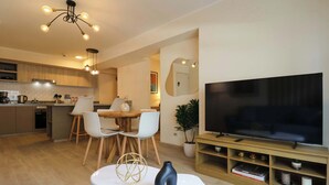 Una cocina moderna con armarios de madera, un comedor con sillas blancas, un televisor sobre un mueble de madera y una planta en maceta.