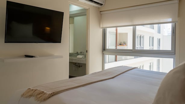 A hotel room with a bed, a TV mounted on the wall, a window with a view of buildings, and a bathroom visible through an open door. - w Comfortable 2BR w Balcony in Miraflores (Lima)