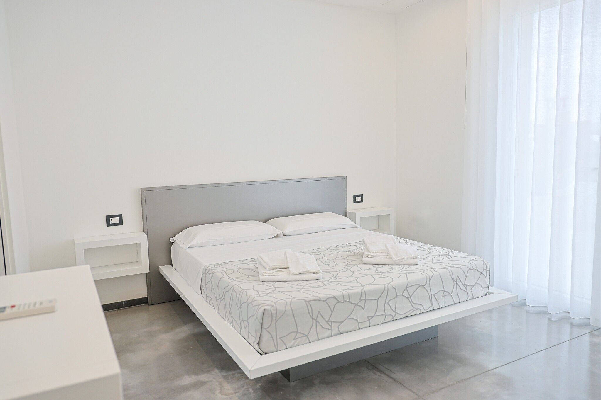 Un dormitorio moderno con cama, mesitas de noche y vistas al exterior a través de cortinas translúcidas.