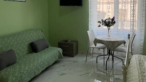 Ein Zimmer mit einem grünen Sofa, einem kleinen Tisch mit Stühlen, einem an der Wand montierten Fernseher und einem Fenster mit gemusterten Vorhängen.
