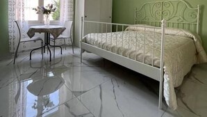 Ein Schlafzimmer mit einem Bett, einem Schreibtisch und einem Stuhl. Es gibt ein Fenster mit Vorhängen, ein Bild an der Wand und eine Klimaanlage.