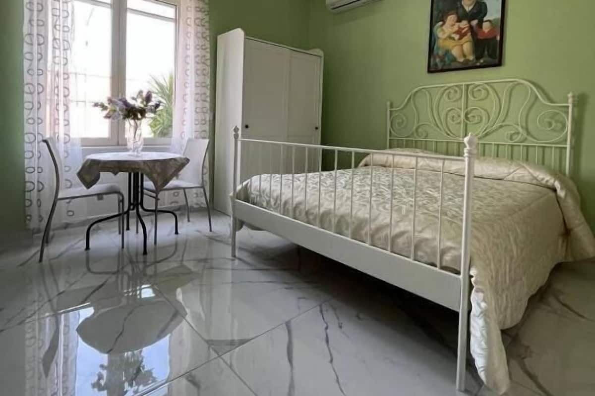 A bedroom with a bed, a desk, and a chair. There is a window with curtains, a painting on the wall, and an air conditioning unit.