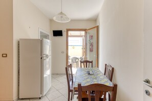 Une cuisine équipée d’un réfrigérateur, d’une table à manger avec des chaises, d’un luminaire suspendu et d’une porte donnant sur un espace extérieur.