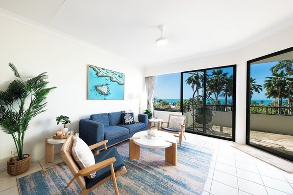 A modern living room with a sofa, armchairs, a coffee table, and a large window offering a view of palm trees and the ocean. - Infinite Blue—Island Views & Coastal Living (Airlie Beach)