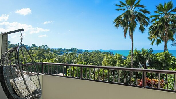 Un balcon agrémenté d’un fauteuil suspendu, offrant une vue panoramique sur des palmiers et une côte lointaine.