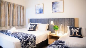 Une chambre d’hôtel avec deux lits, une grande fenêtre avec des rideaux, une table de chevet avec une lampe et deux tableaux encadrés au mur.