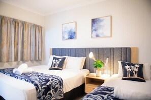 A hotel room with two beds, a large window with curtains, a nightstand with a lamp, and two framed pictures on the wall. - Infinite Blue (Airlie Beach)