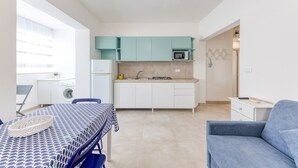 A compact kitchen with teal cabinets, a microwave, and a sink. There is a dining table with a patterned tablecloth and a blue chair.
