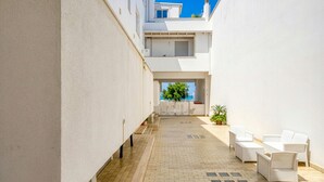 A narrow courtyard with a tiled floor, white walls, and a seating area with white wicker furniture.