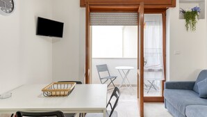Een kamer met een eettafel, stoelen, een bank, een televisie, een schuifdeur naar een balkon en een klok.