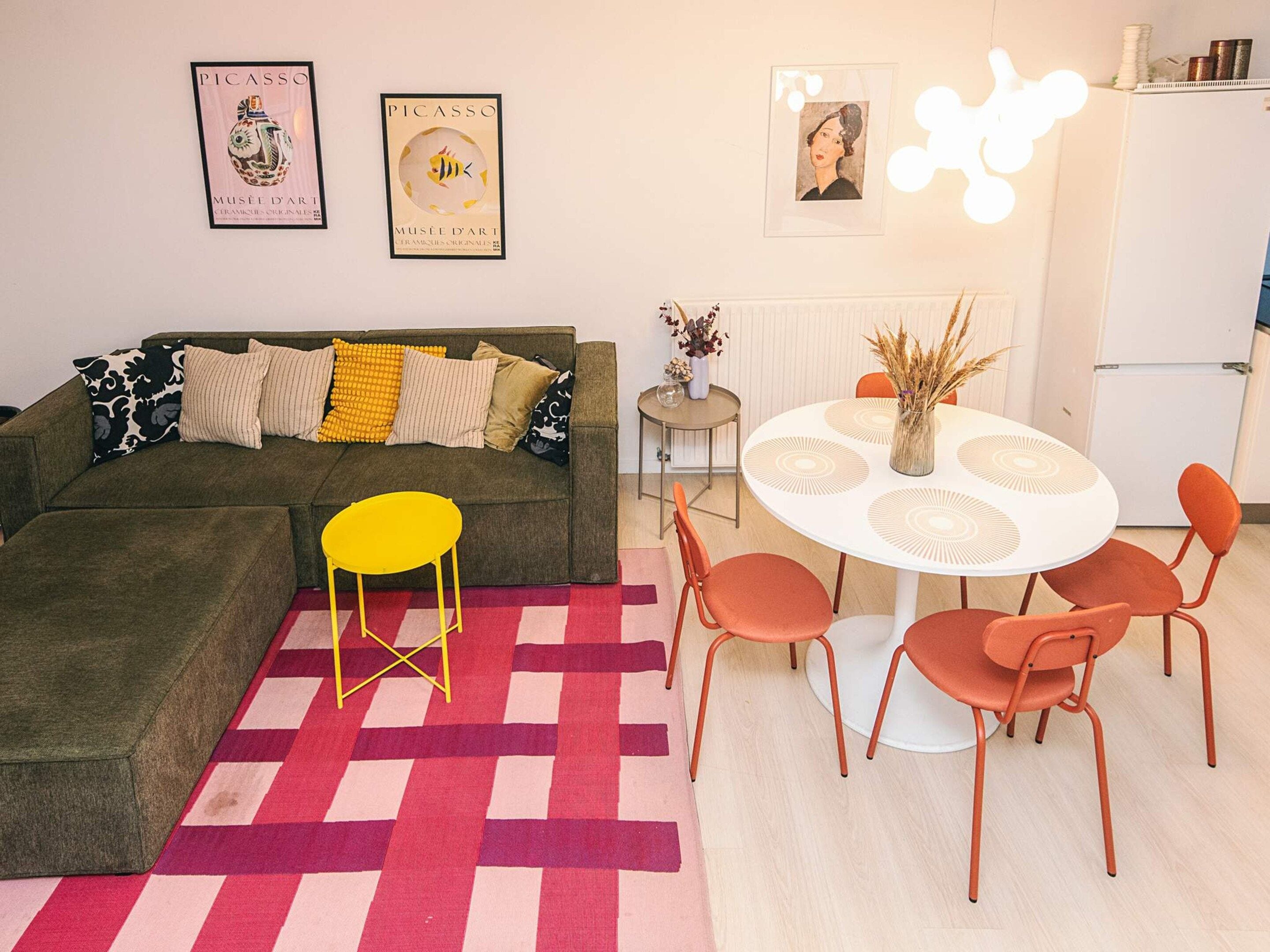 Una sala de estar con un sofá, una silla amarilla, una mesa de comedor con sillas naranjas y un tapete estampado.