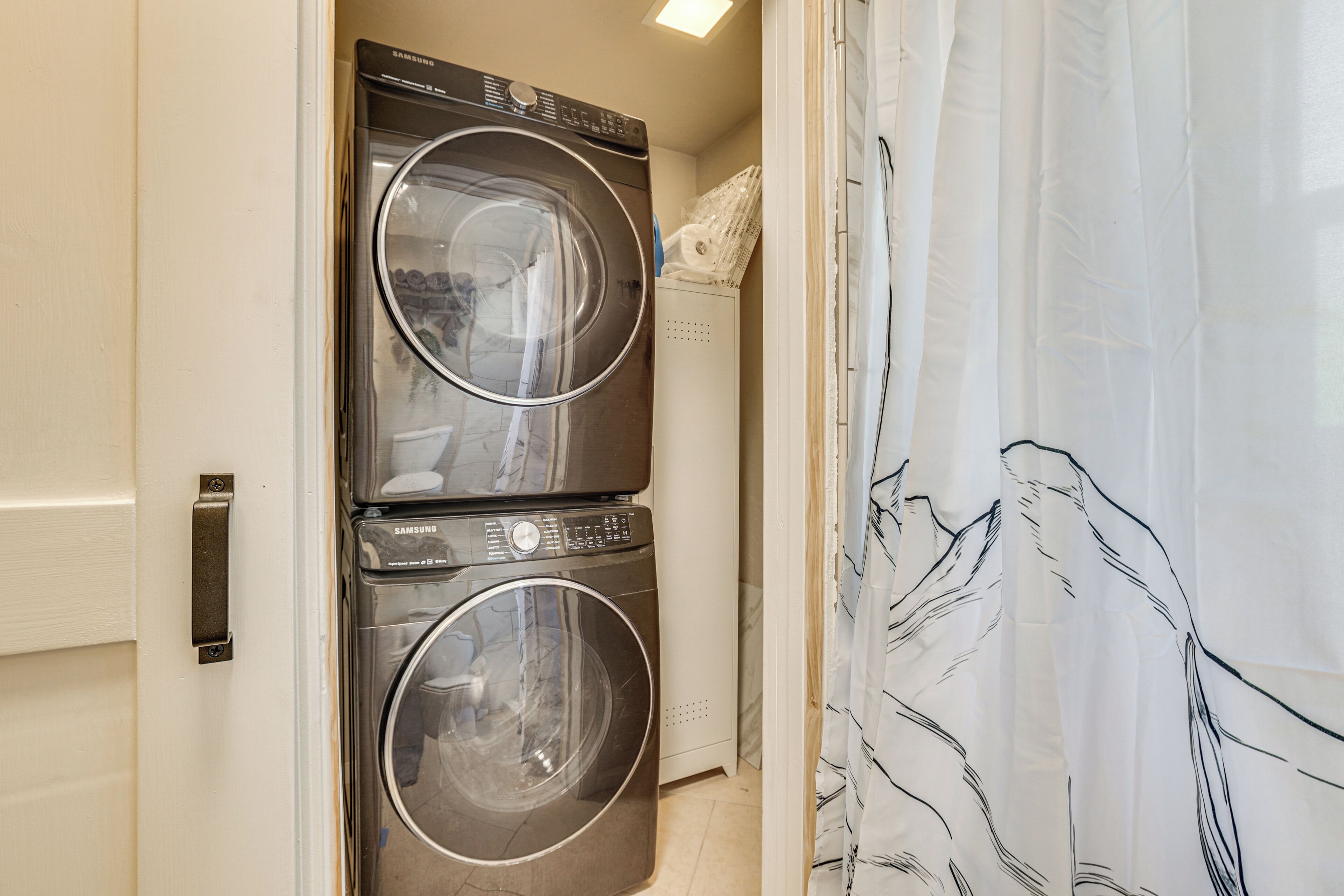 A laundry area with two stacked Samsung washing machines, a white dryer, and a curtain with a leaf pattern.