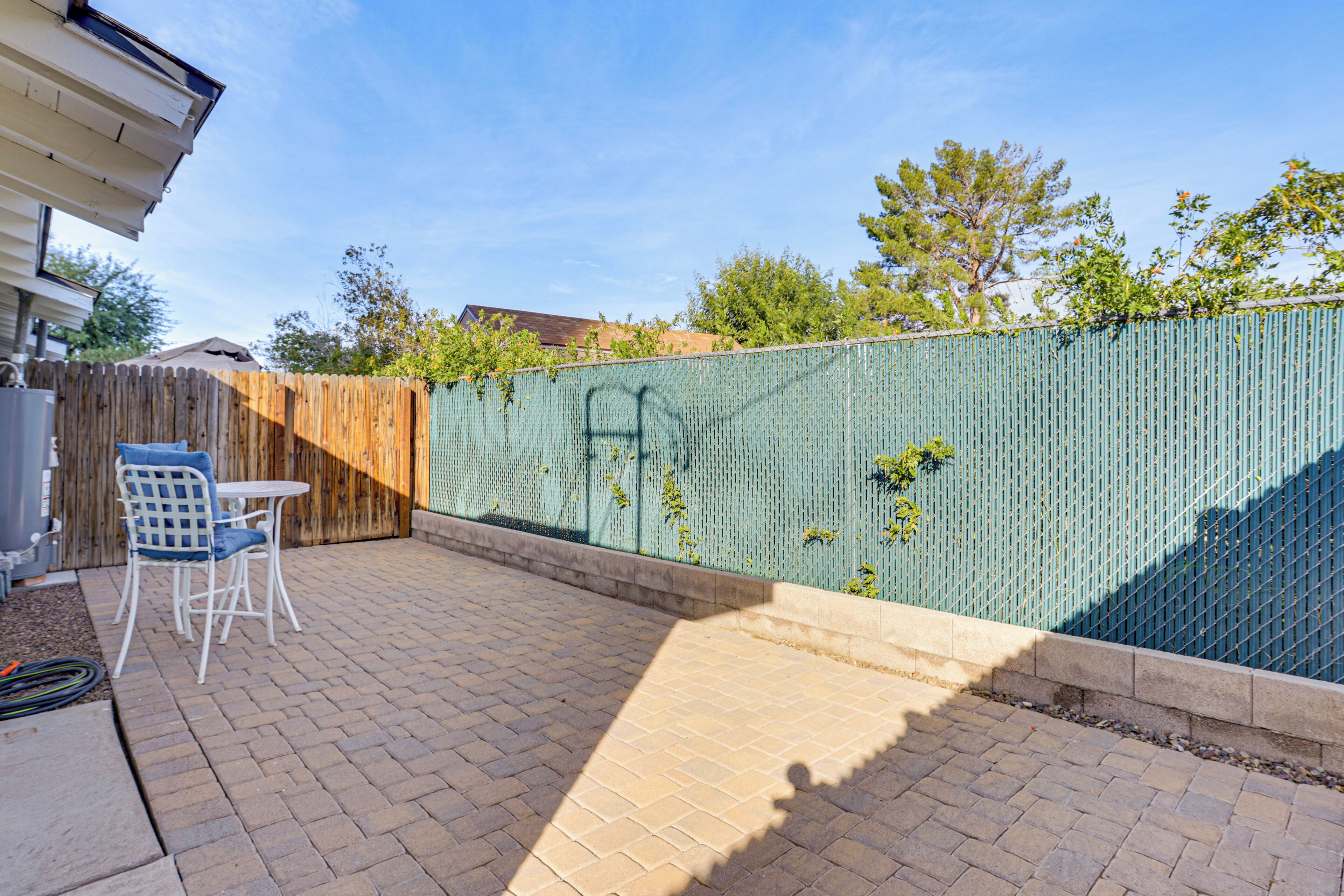 A patio area with a brick floor, a white table and chair, and a green fence with some foliage.
