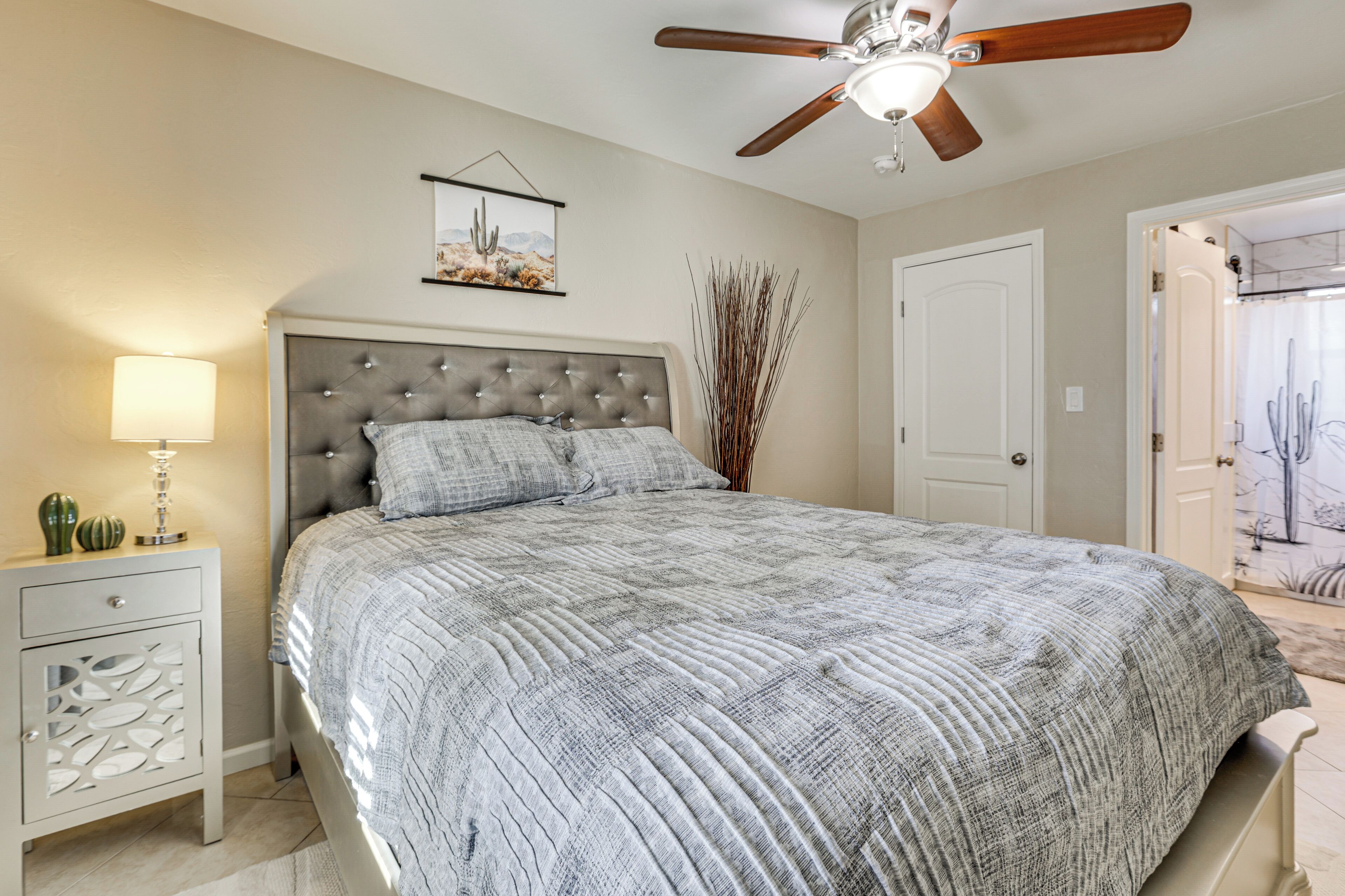 A bedroom with a tufted headboard, a bed with a patterned bedspread, a bedside table with a lamp, a ceiling fan, and a framed picture on the wall.