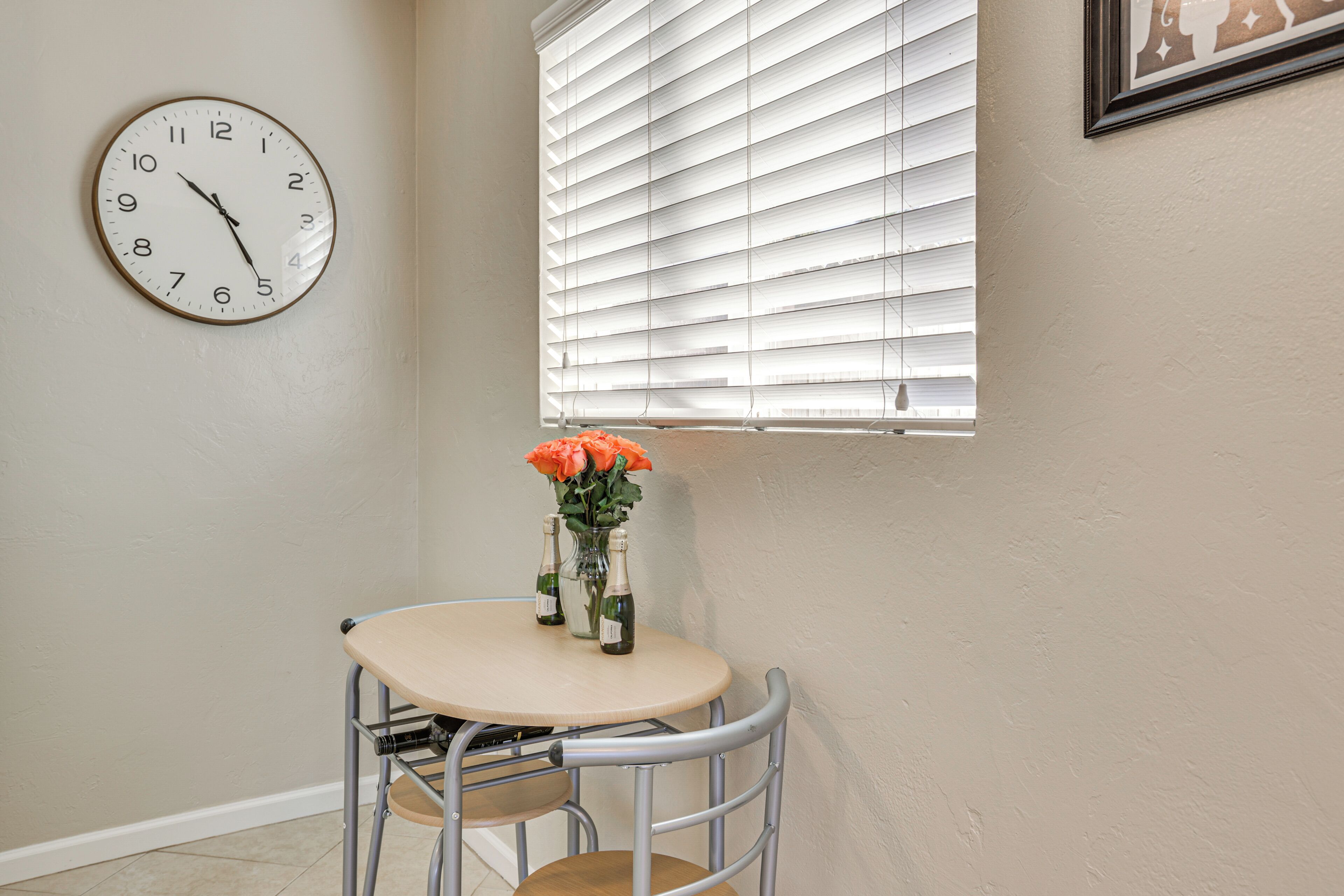 A room with a round table, a metal chair, a wall clock, a window with blinds, and a vase with flowers.