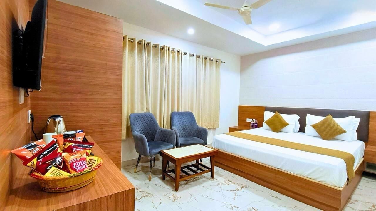 A hotel room with a bed, two armchairs, a small table, a TV, and a basket of snacks.
