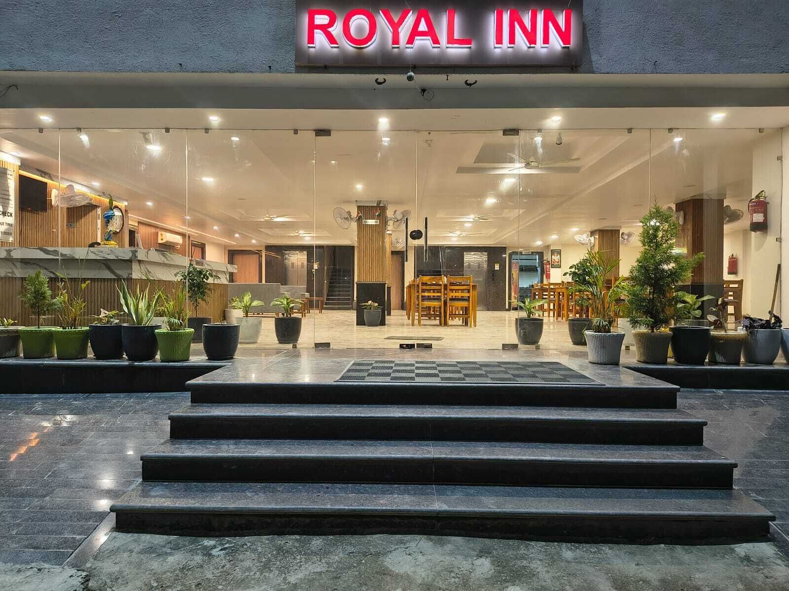 The exterior of the Royal Inn with a modern design, featuring a glass facade, a well-lit entrance, and a welcoming reception area with potted plants.