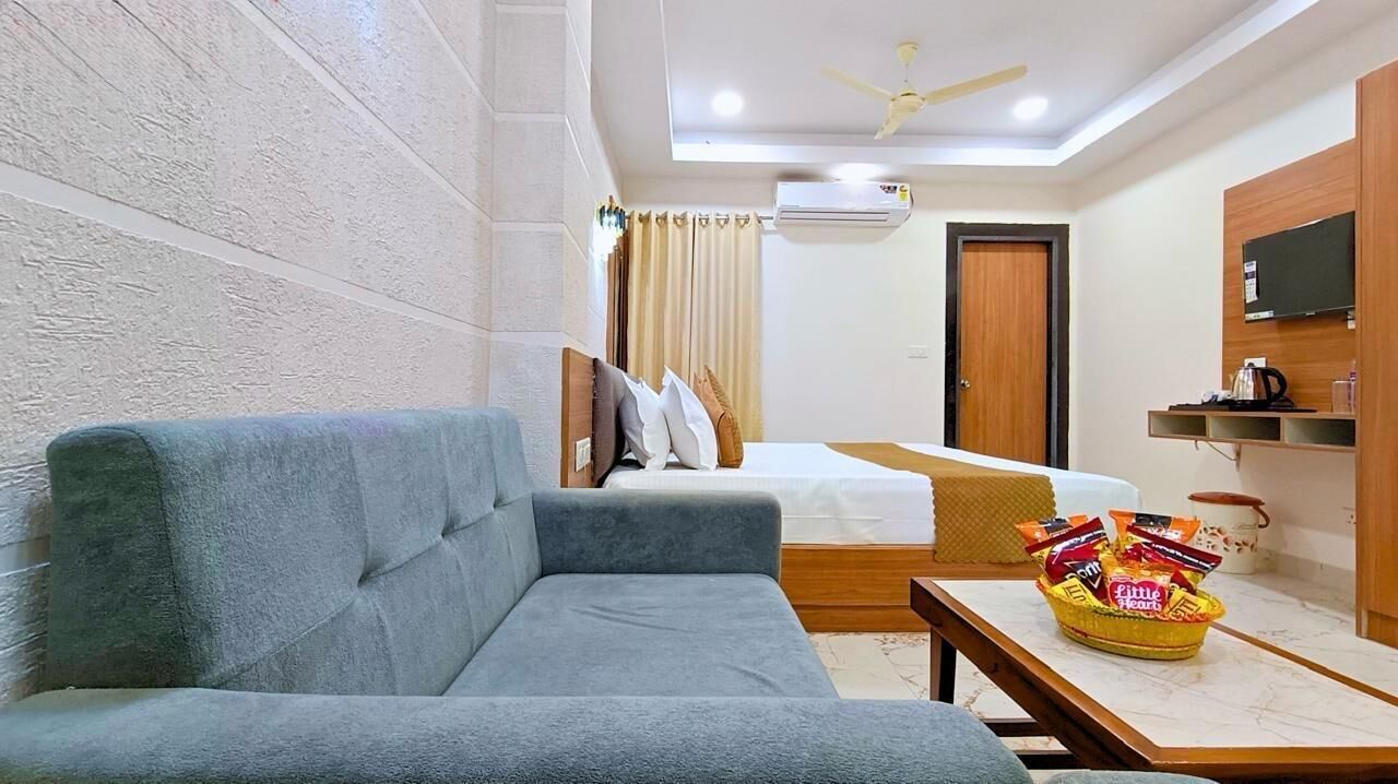 Une chambre d’hôtel avec un canapé gris, un lit avec une literie blanche, une petite table avec des collations, un téléviseur posé sur une étagère en bois et un climatiseur fixé au mur.