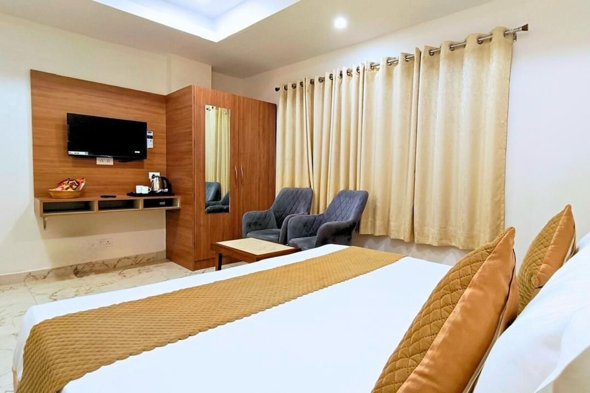 A hotel room with a bed, a TV mounted on the wall, a seating area with chairs, and a window with curtains.