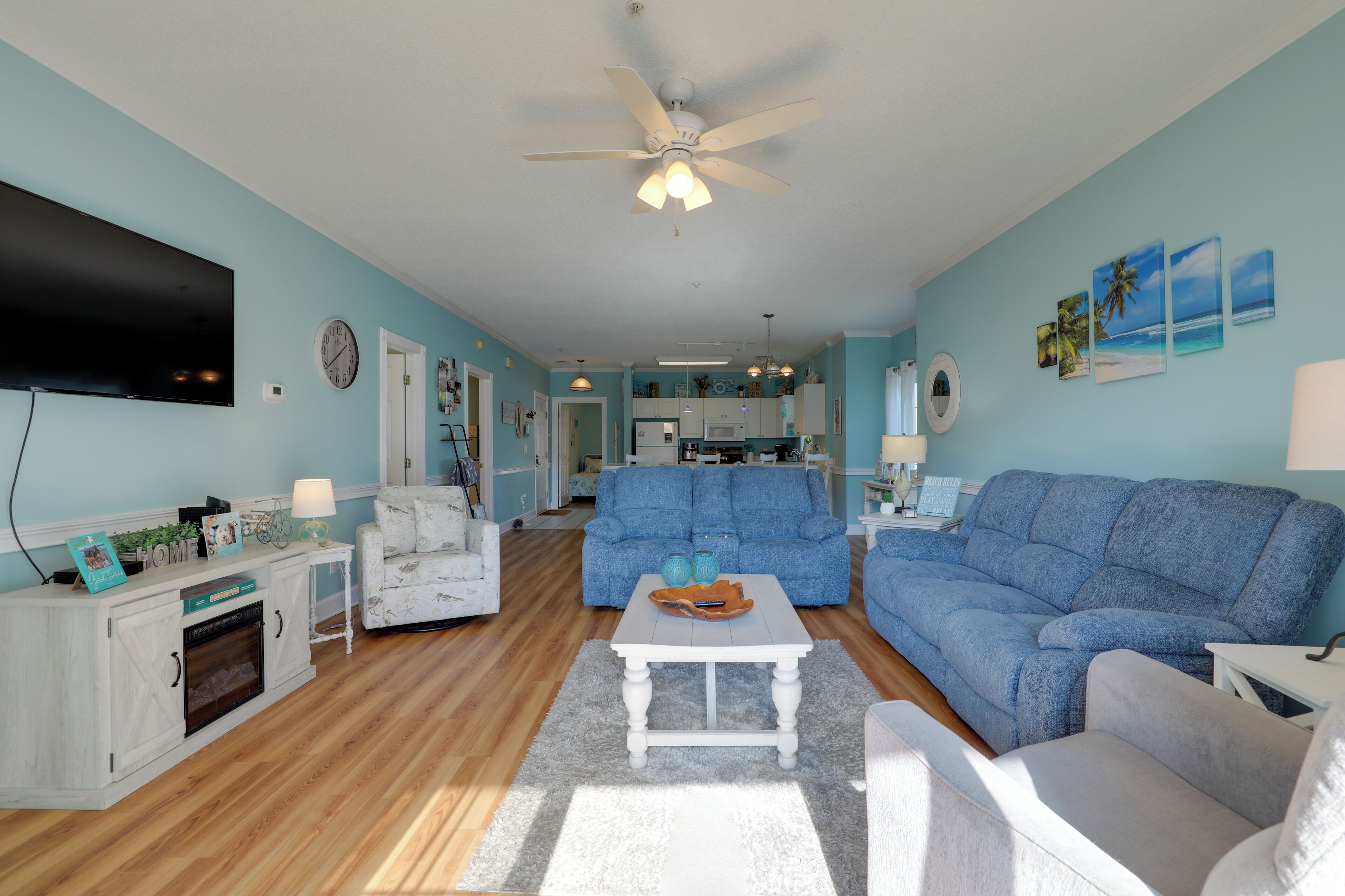 A living room with a blue sofa, white armchairs, a coffee table, and a TV. There is a kitchen in the background.