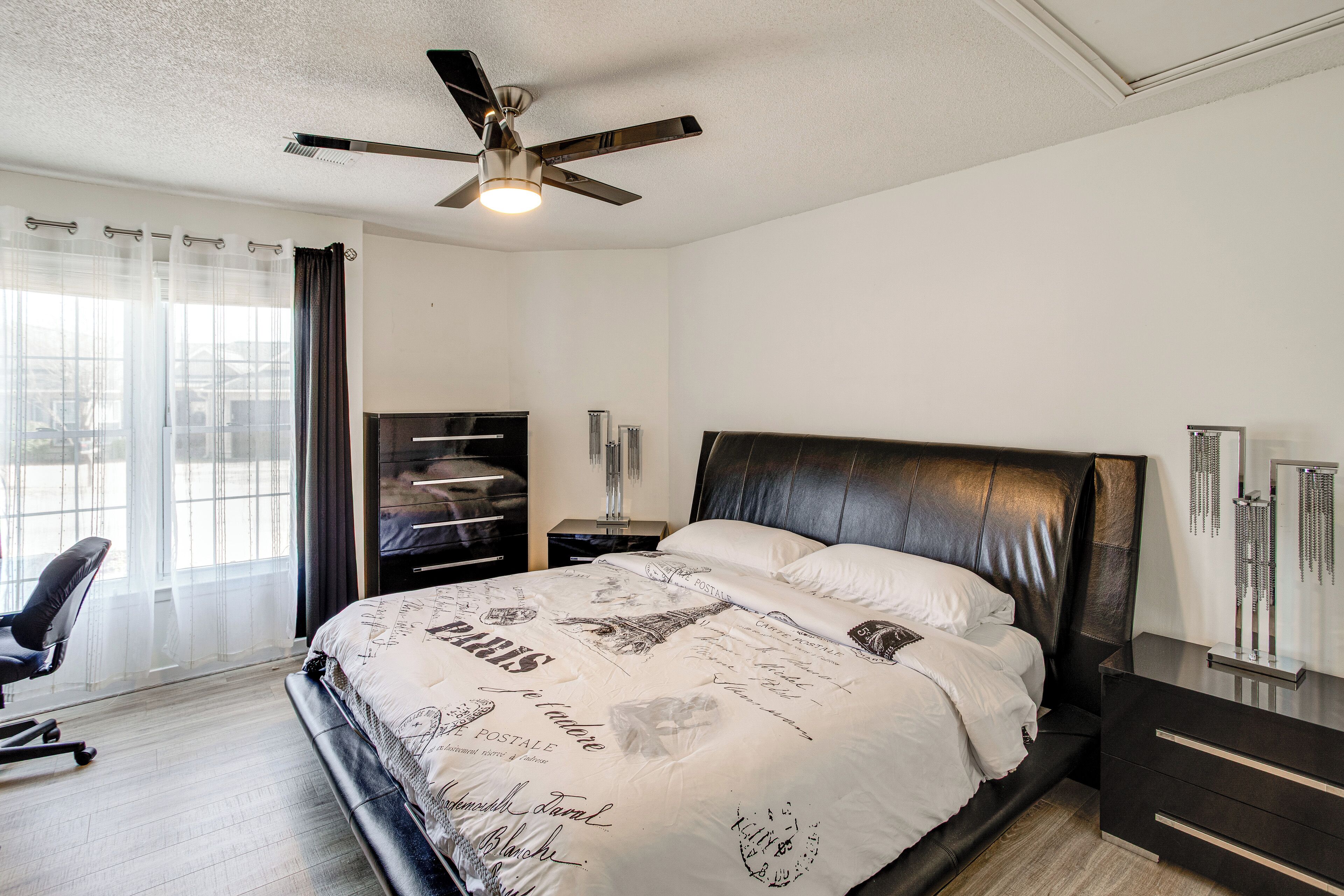 A bedroom with a bed, a desk, a chair, a ceiling fan, and a window with curtains.